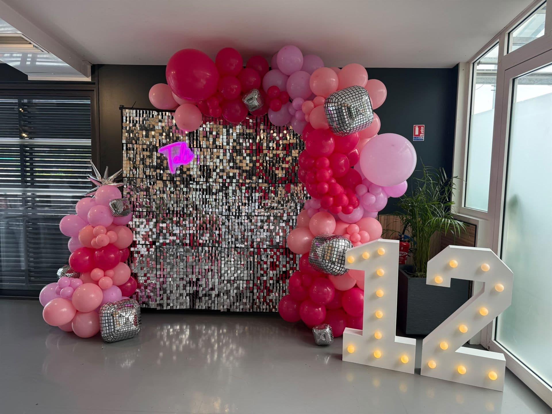 Décor de Bat Mitsva : mur à sequins argentés, prénom en néon rose, arche de ballons roses et fuchsia avec boules à facettes et chiffres lumineux 12