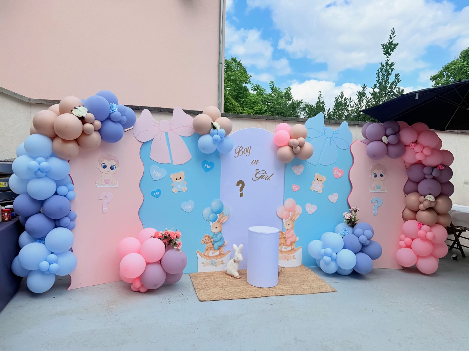Décor de gender reveal « Boy or Girl » en terrasse : panneaux roses et bleus avec nœuds, cœurs He et She, lapins et arches de ballons pastel